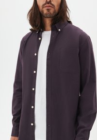 Man med axellångt hår och skägg som bär en mörklila skjorta med knappar över en vit t-shirt mot en enhetlig bakgrund.