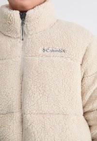 Fleece jacket in light beige with a high collar, featuring a front zipper and the Columbia logo in gray on the chest. Soft, textured fabric.