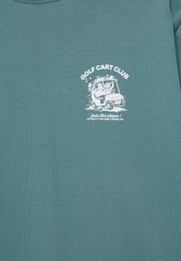 Graphique blanche sur tissu vert montrant deux personnes conduisant joyeusement une voiturette de golf avec des clubs de golf, texte : Golf Cart Club, Rejoignez le chaos.
