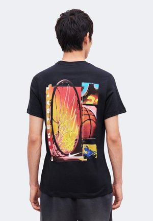 Mand iført sort t-shirt med farverigt basketball-tema grafik, der viser kurv, bold, trøje og sneaker på bagsiden.