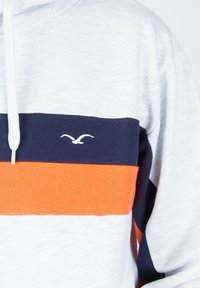 Mikina s vodorovnými námořnickými modrými a oranžovými pruhy přes hrudník, s bílým logem racka na námořnickém pruhu.