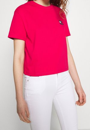 Kobieta ubrana w jaskrawoczerwony krótki krótki t-shirt z małym logo na piersi i białe dopasowane spodnie na jednolitym tle.