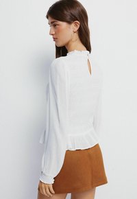 Bílá dlouhá blůza s texturovanou tkaninou, řaseným výstřihem a detaily klíčové dírky na zádech, spárovaná s hnědou semišovou mini sukní.