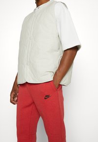 Ljusgrå vadderad väst med rund sömnad, med framknappar och krage, bärs över en vit skjorta tillsammans med röda sweatpants.