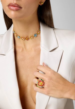 Vrouw draagt goudkleurige sieradenset met bloemen, versierd met rode, blauwe en gele emaille bloemen: halsketting, oorbellen en een grote ring in bloemvorm.