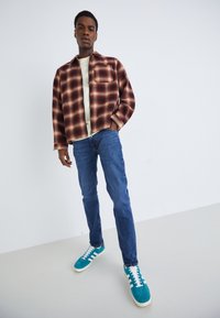 Camisa a cuadros en tonos rojos y marrones, camiseta blanca debajo, jeans de mezclilla azul y zapatillas de deporte en color azul verdoso con acentos blancos.