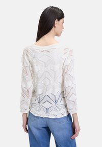 Maglione bianco a maglia con motivo geometrico in pizzo, caratterizzato da maniche lunghe e un ampio scollo, abbinato a jeans azzurri chiari.