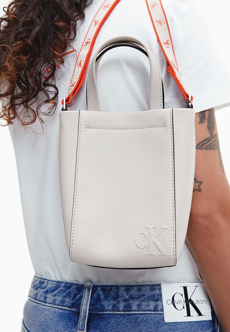 Šedá kabelka z umělé kůže s hladkým povrchem, obdélníkovým tvarem, horními uchy a nastavitelným oranžovým popruhem. Má embosované logo.