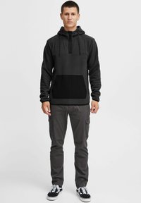 Zwarte en donkergrijze hoodie met een halve rits, voorzak, en contrasterende texturen; gecombineerd met grijze cargobroeken en zwarte sneakers.