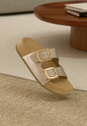 Sandale en toile beige avec deux boucles dorées ajustables reposant sur un tapis tissé texturé à côté d'une table ronde en bois avec des livres.