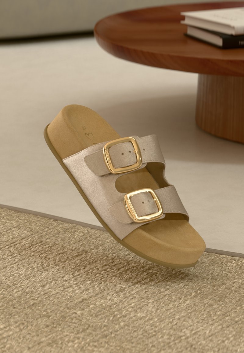 Sandale en toile beige avec deux boucles dorées ajustables reposant sur un tapis tissé texturé à côté d'une table ronde en bois avec des livres.