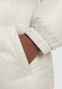 Chaqueta de plumas crema con un acabado brillante, que presenta puños elásticos y un bolsillo lateral. Logo "THERMA-FIT" bordado en la manga.