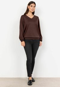 Suéter marrón de manga larga y cuello en V con un ajuste relajado, combinado con jeans negros ajustados y mocasines negros, frente a un fondo neutral.