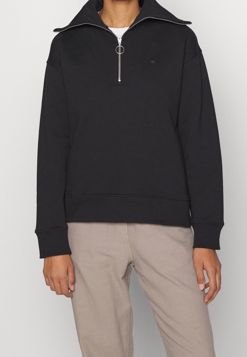 Personne portant un pull noir à demi-fermeture éclair avec une tirette en anneau et un pantalon beige sur un fond uni.