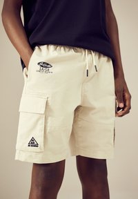 Light beige cargo shorts with an elastic waistband, side pockets, and a triangular patch reading "No Risk No Reward." Adjustable drawstring details.
