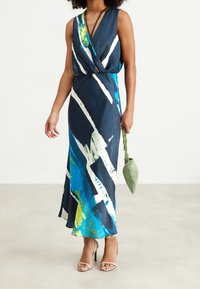 Maxi šaty v námořnické barvě s abstraktními bílými a modrými vzory, s překlápěcím designem a výstřihem do V. Doplněné o zelenou texturovanou kabelku.