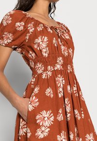 Robe florale brune avec des manches bouffantes, taille smockée et poches latérales, présentant des motifs de fleurs blanches et rose pâle. Tissu doux et texturé.