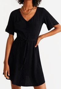 Vestido negro con escote en V y manga corta, con un cierre frontal de botones y cintura ajustada con lazo. Textura suave y corte holgado.