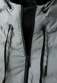 Vadderad jacka i ljusgrått med svart dragkedja och snoddar. Har en hög krage och präglad varumärkesetikett på placket.