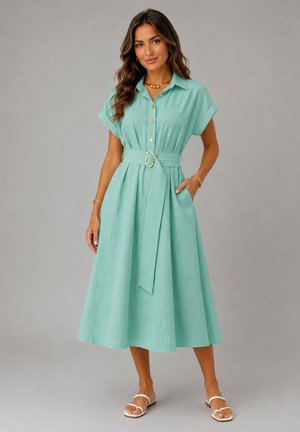 Femme portant une robe midi ceinturée bleu sarcelle clair avec des manches courtes et des sandales blanches, debout devant un fond gris uni.