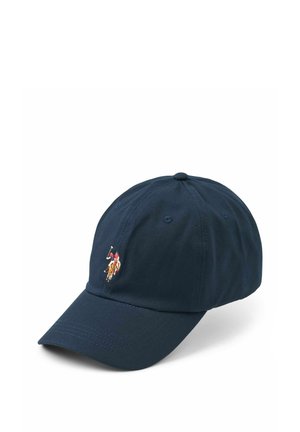 Glimlachende vrouw met lang blond haar, die een marineblauwe pet met logo, een zwart V-hals shirt en een lichtpaarse doorgestikte jas draagt tegen een effen achtergrond.