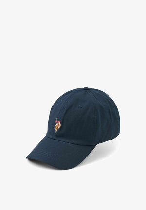 Smilende kvinde med langt blondt hår iført en marineblå kasket med logo, sort V-hals trøje og let lilla quiltet jakke foran en ensfarvet baggrund.