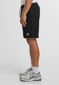 Černé bavlněné šortky s elastickým páskem a bočními kapsami, s malým bílým logem na levé noze. Nosené se stříbrnými sportovními botami.