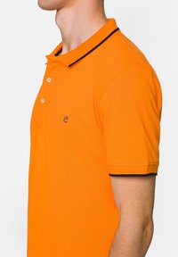 Pomarańczowa koszulka polo z kołnierzykiem i trzema guzikami. Czarny akcent na kołnierzyku i ściągaczach rękawów. Dyskretny detal logo na klatce piersiowej.