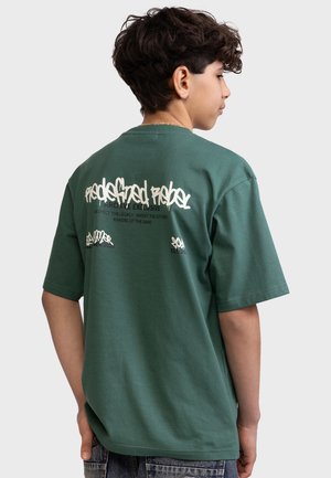 Teenagedreng med krøllet mørkt hår iført en grøn oversized T-shirt med hvid graffiti-stil tekst på ryggen, stående foran en ensfarvet baggrund.