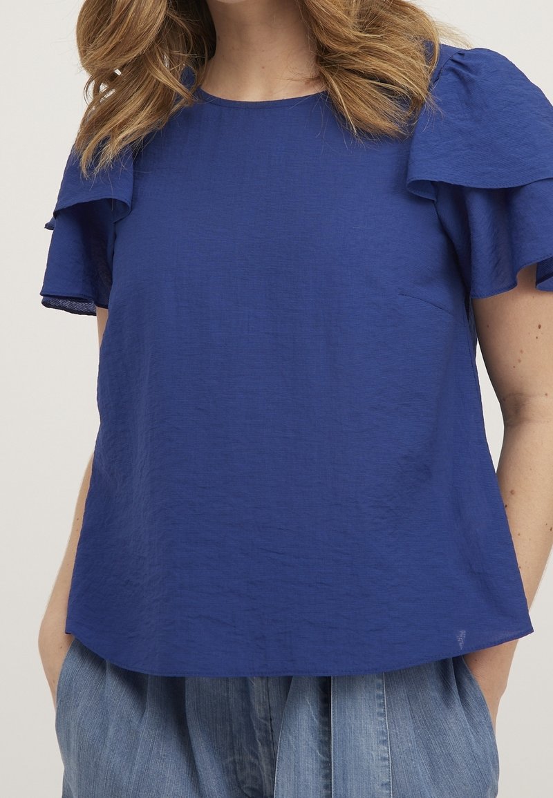 Femme portant un chemisier bleu à manches courtes avec des manches volantées et un pantalon ample bleu clair, debout les mains dans les poches.