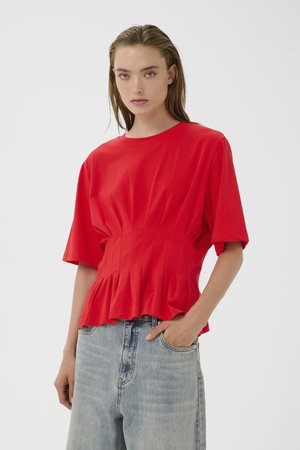 Frau mit nassen Haaren, die ein rotes Kurzarmoberteil mit Falten und hellblaue hoch taillierte Jeans trägt, Hand in der Tasche.