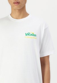 Bílý bavlněný tričko s potiskem zeleného a žlutého textu na levé části hrudníku s nápisem "Užijte si dovolenou" a "The Easy Peasy Company."