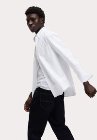 Weißes Hemd mit langen Ärmeln, einer Brusttasche, über einem grauen Shirt und dunklen Jeans mit kontrastierenden Nähten getragen.