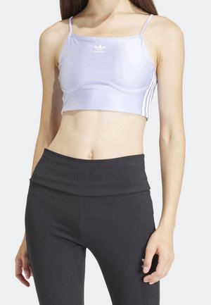 Kobieta nosząca jasnofioletowy krótki top Adidas i czarne legginsy z wysokim stanem na białym tle.