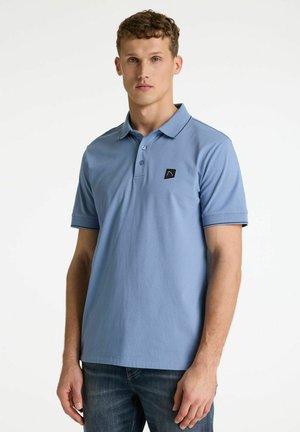 Jonge man die een lichtblauw poloshirt met korte mouwen draagt, met een klein zwart vierkant logo op de borst, gecombineerd met een donkere spijkerbroek.
