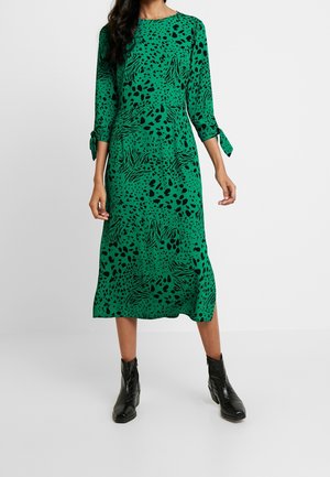 Groene jurk van gemiddelde lengte met zwart dierenprintpatroon. Heeft lange mouwen met strikkoorden en een ronde halslijn. Gecombineerd met zwarte enkellaarsjes.