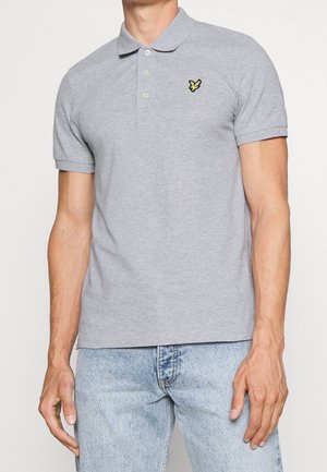 Mand iført en lysegrå kortærmet polo med en gul fuglelogo på brystet og lyseblå denimjeans.