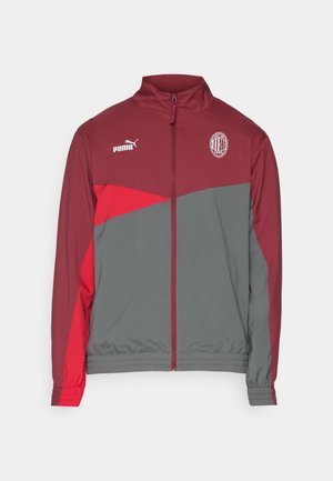 Giacca zip-up rossa e grigia con colletto alto. Presenta il logo Puma e un emblema della squadra sul petto. Polsini ed elastico in vita. Tessuto morbido.