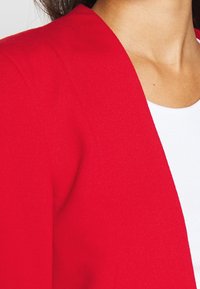 Punainen neulecardigan, jossa on v-pääntie ja pehmeä tekstuuri, joka peittää osittain valkoista yläosaa. Saumayksityiskohdat näkyvät hartioilla.