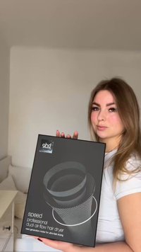 Frau mit langen Haaren hält eine schwarze Box mit der Aufschrift „ghd speed professional dual air-flow hair dryer“ in einem hellen Raum.