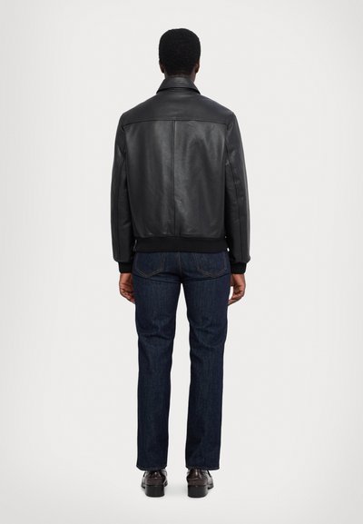 Chaqueta de cuero negra con un dobladillo acanalado, detalle de yugo trasero y dos bolsillos laterales, combinada con jeans de mezclilla oscura y zapatos negros pulidos.