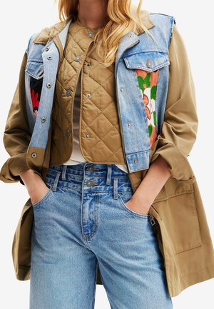 Femme portant plusieurs couches de vêtements : gilet matelassé beige sous un gilet en jean bleu avec des panneaux floraux, veste beige, et jean taille haute bleu clair, mains dans les poches.