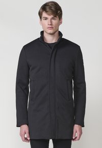 Abrigo exterior negro con cuello alto, cierre de cremallera y dos bolsillos frontales. Fabricado en un tejido suave con un corte ajustado.