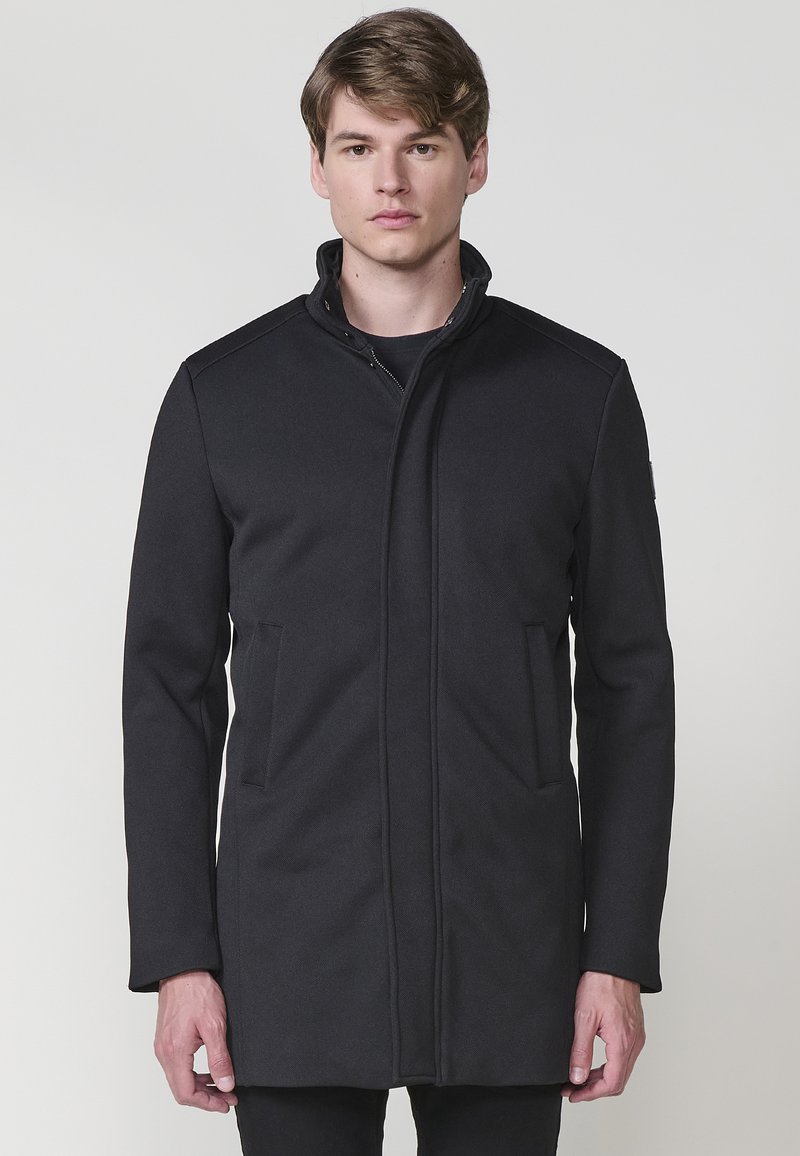 Abrigo exterior negro con cuello alto, cierre de cremallera y dos bolsillos frontales. Fabricado en un tejido suave con un corte ajustado.