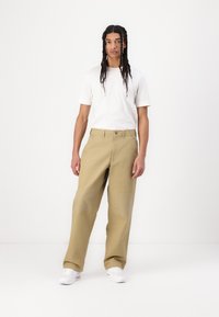 Beige vida byxor med texturerade detaljer på knäna, tillsammans med en vit t-shirt med kort ärm och vita sneakers.