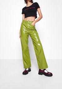 Pantalones de vinilo verdes, de cintura alta y brillantes, con un corte suelto, combinados con una camiseta negra recortada con texto rojo. Unos zapatos burdeos gruesos completan el look.