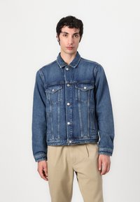 Denimjakke i middels blå, med krage, to lommer foran med klaffer, og slitte detaljer på ermer og kant.