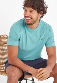 Man with curly hair wearing a turquoise T-shirt and navy shorts sitting on a wicker chair, smiling and looking right.