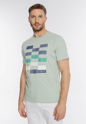 Mężczyzna z krótkimi włosami i brodą, ubrany w jasnografitowy t-shirt z nadrukiem i białe spodnie, stojący na jednolitym białym tle.