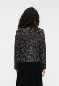 Veste noire texturée avec des éclaboussures colorées, manches longues et col arrondi, associée à une jupe noire plissée.